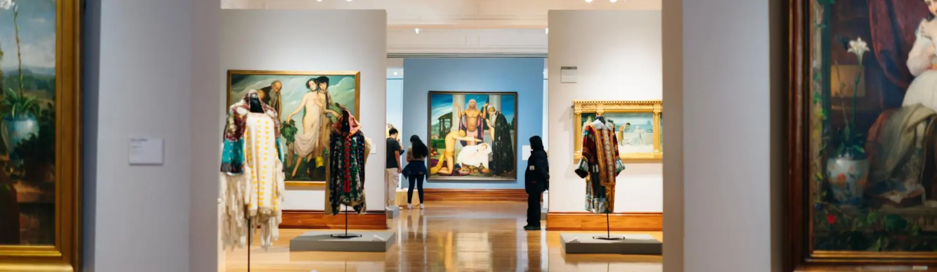 Ausstellungssaal eines Museums mit Gemälden und aufgestellten Kostümen, Besucher betrachten die Werke